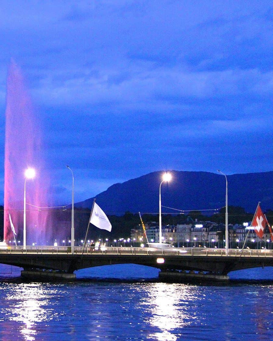 The Geneva Jet d'eau lit up in the evening above the harbour, with the lights of the quays reflected in the lake