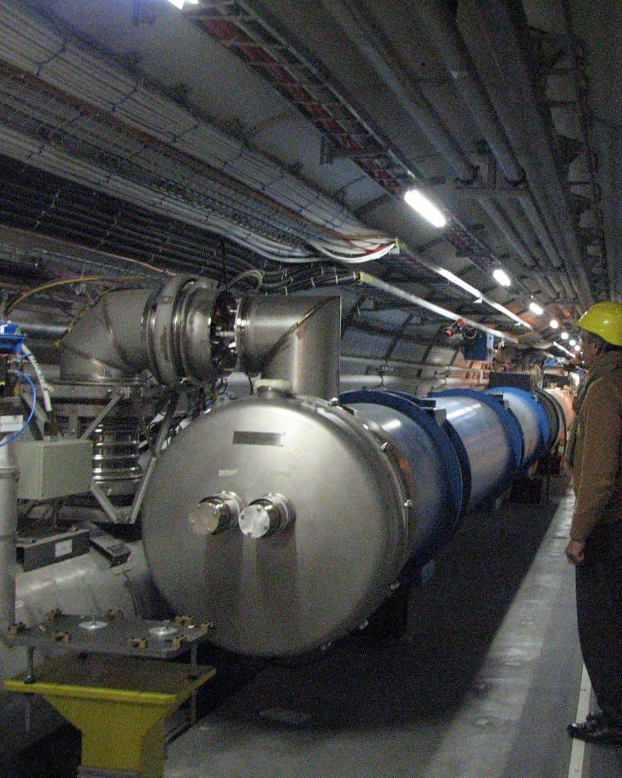 Le tunnel du LHC près du point 5, avec ses aimants supraconducteurs bleus alignés et un technicien casqué, une zone fermée au public pendant le LS3